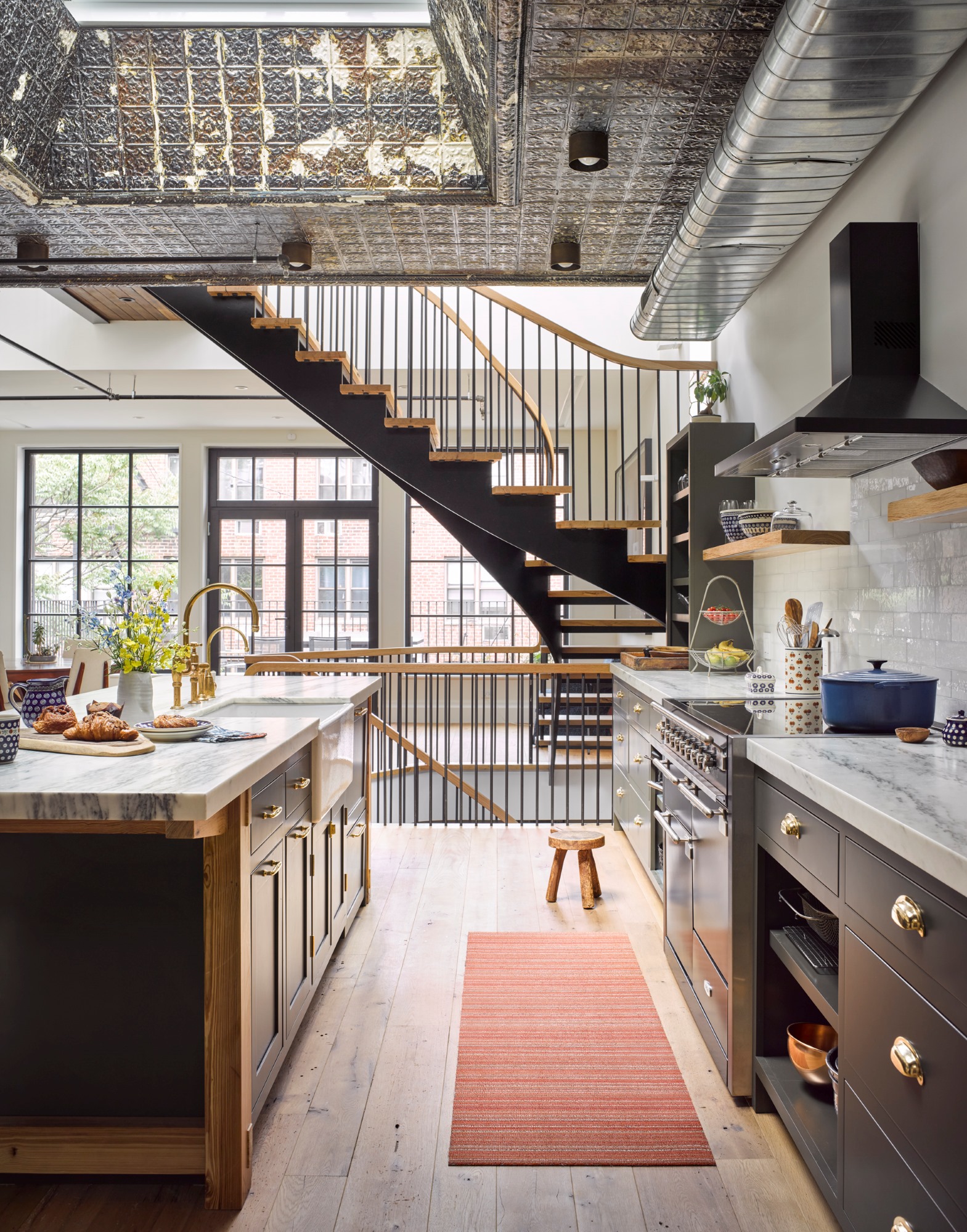 Kitchen in a New York City apartment. The kitchen has wide plank wood floors, white marble countertops, and reclaimed tin ceilings. Beyond the kitchen, the rear of the apartment and living room can be seen through an open, sculptural staircasae.