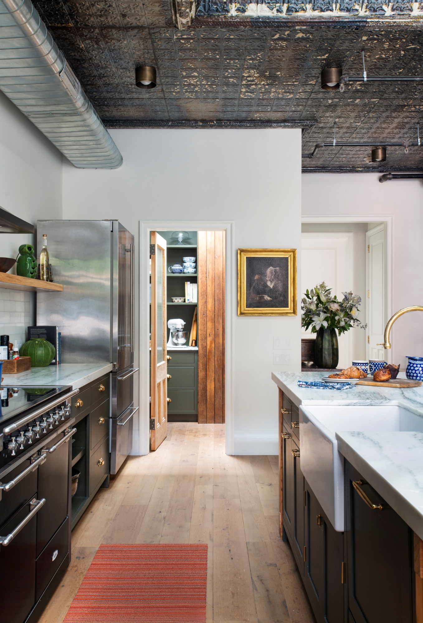 Kitchen in a New York City apartment. The kitchen has wide plank wood floors, white marble countertops, and reclaimed tin ceilings. The reclaimed pantry door opens into the large pantry. Warm wood tones and green painted millwork match the kitchen.