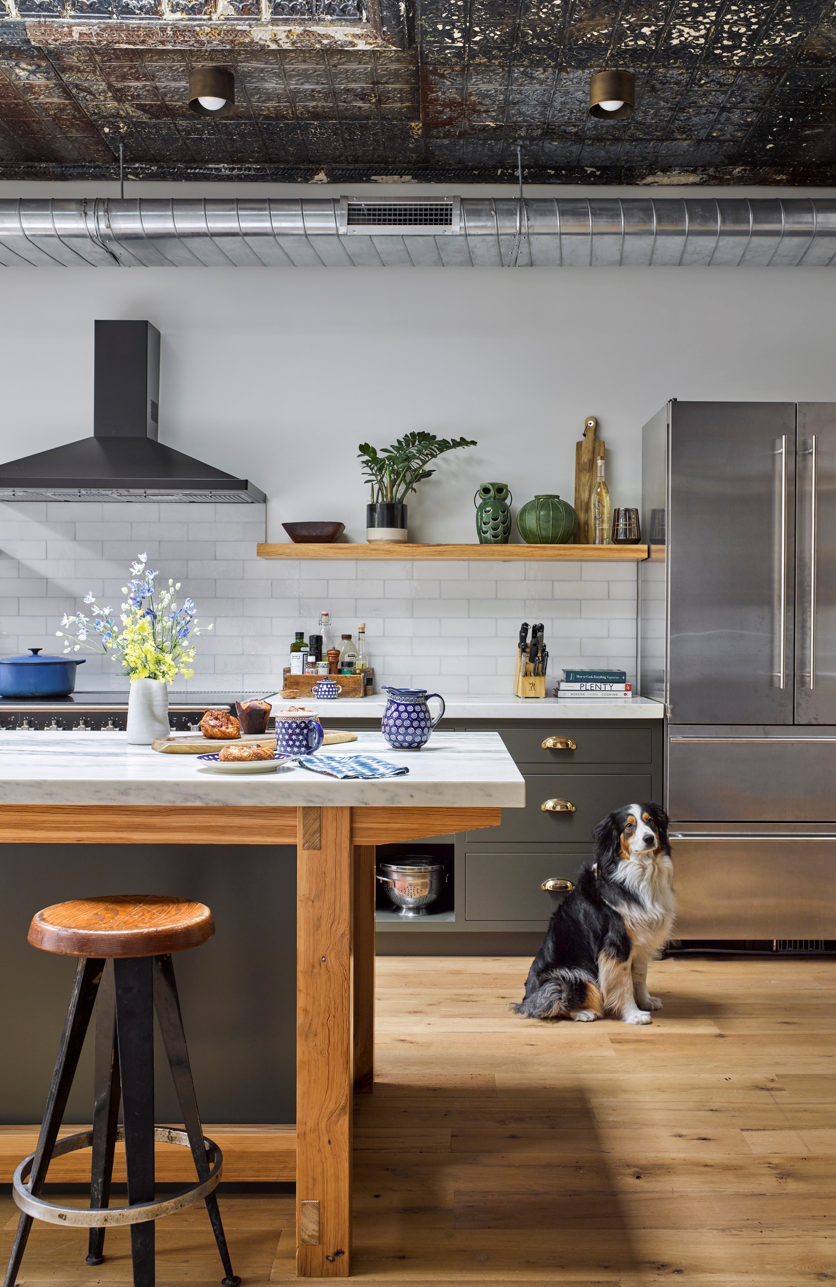 Kitchen in a New York City apartment. The kitchen has wide plank wood floors, white marble countertops, and reclaimed tin ceilings. On the counter, pastries and coffee mugs are laid out next to a floral arrangement. A dog sits in front of a stainless steel refrigerator at the right of the frame.