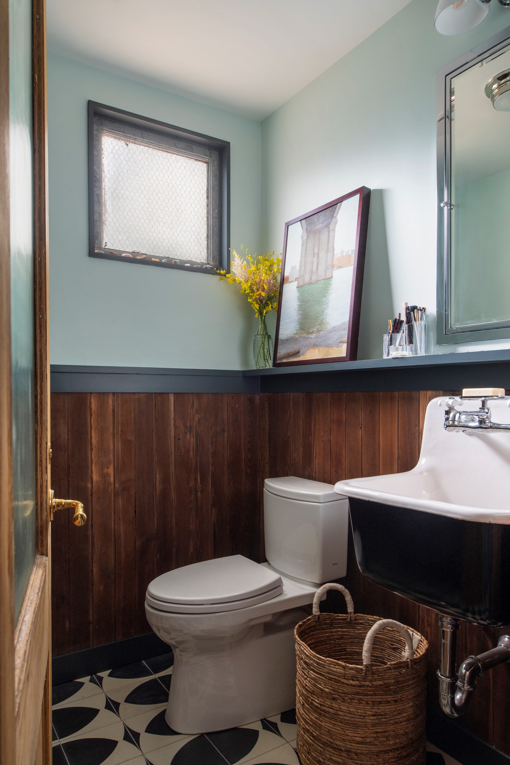 Powder room in a Manhattan apartment. At the left of the frame, the reclaimed wood door with brass hardware can be seen. The floors are black and white tile, and the bottom half of the walls are clad in reclaimed wood. The top half of the walls are covered in a light seafoam green color.