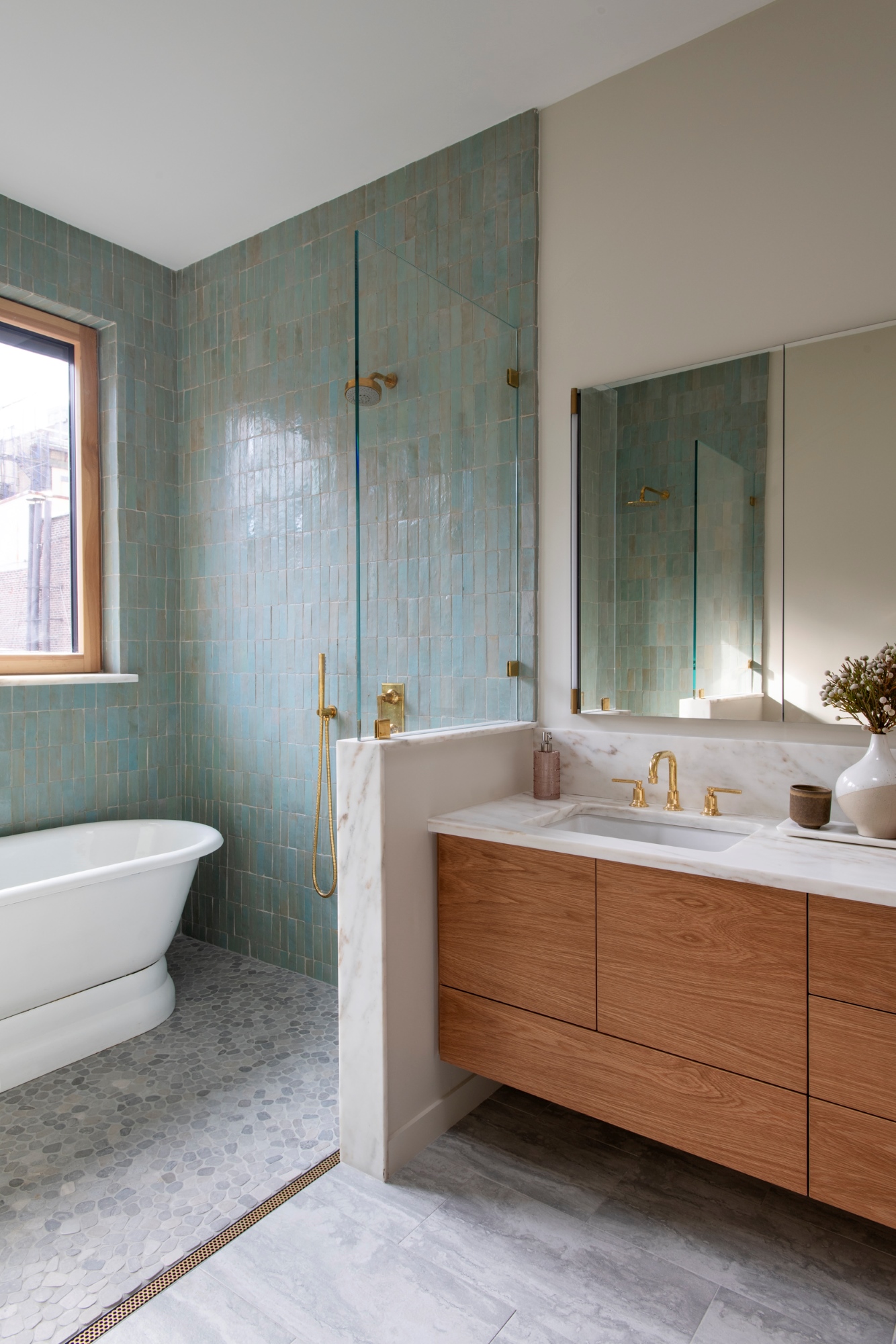 Primary bathroom in a Manhattan apartment. The shower stall, on the left side of the image, has light blue and green tiled walls, gold hardware, round grey floor tiles, and a reclaimed freestanding tub. The vanity area has white walls, grey stone flooring, a white vanity countertop and a floating wood vanity.