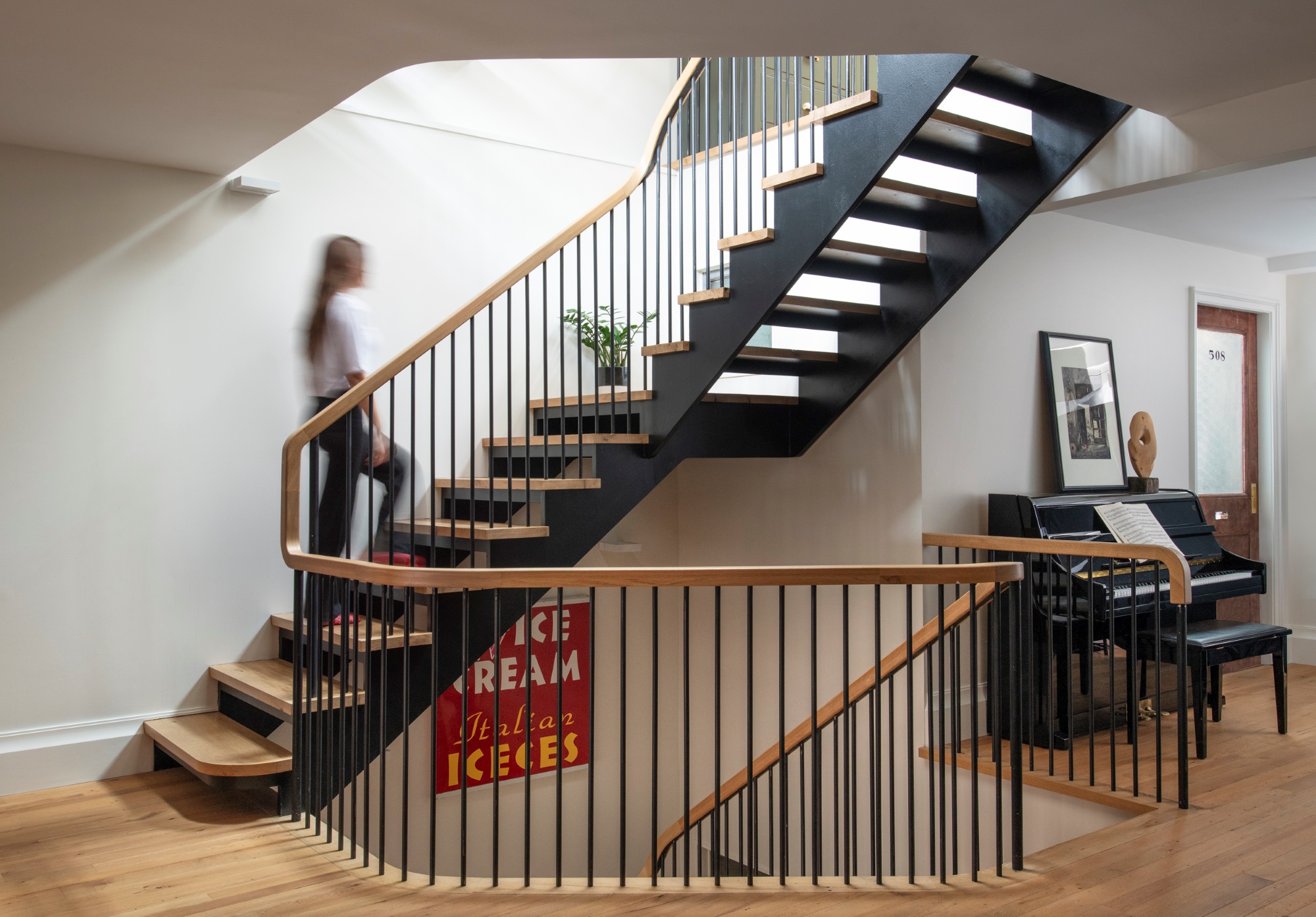 Open staircase with natural wood risers and handrail, and black metal stringers and balusters. The staircase is open to the floors above and below. A woman with a white shirt and black pants walks up the staircase. To the right of the staircase, an upright piano has artwork on top of it, and a reclaimed door leads to another room of the apartment.