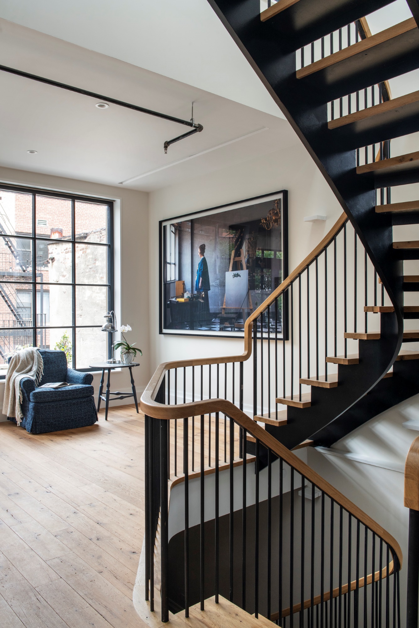 The foreground of the image contains a black metal and natural wood, open-riser, open-stringer sculptural staircase. Beyond the staircase, an armchair sits in front of a window. Hanging on the wall next to the staircase is a photograph that references a Vermeer painting.