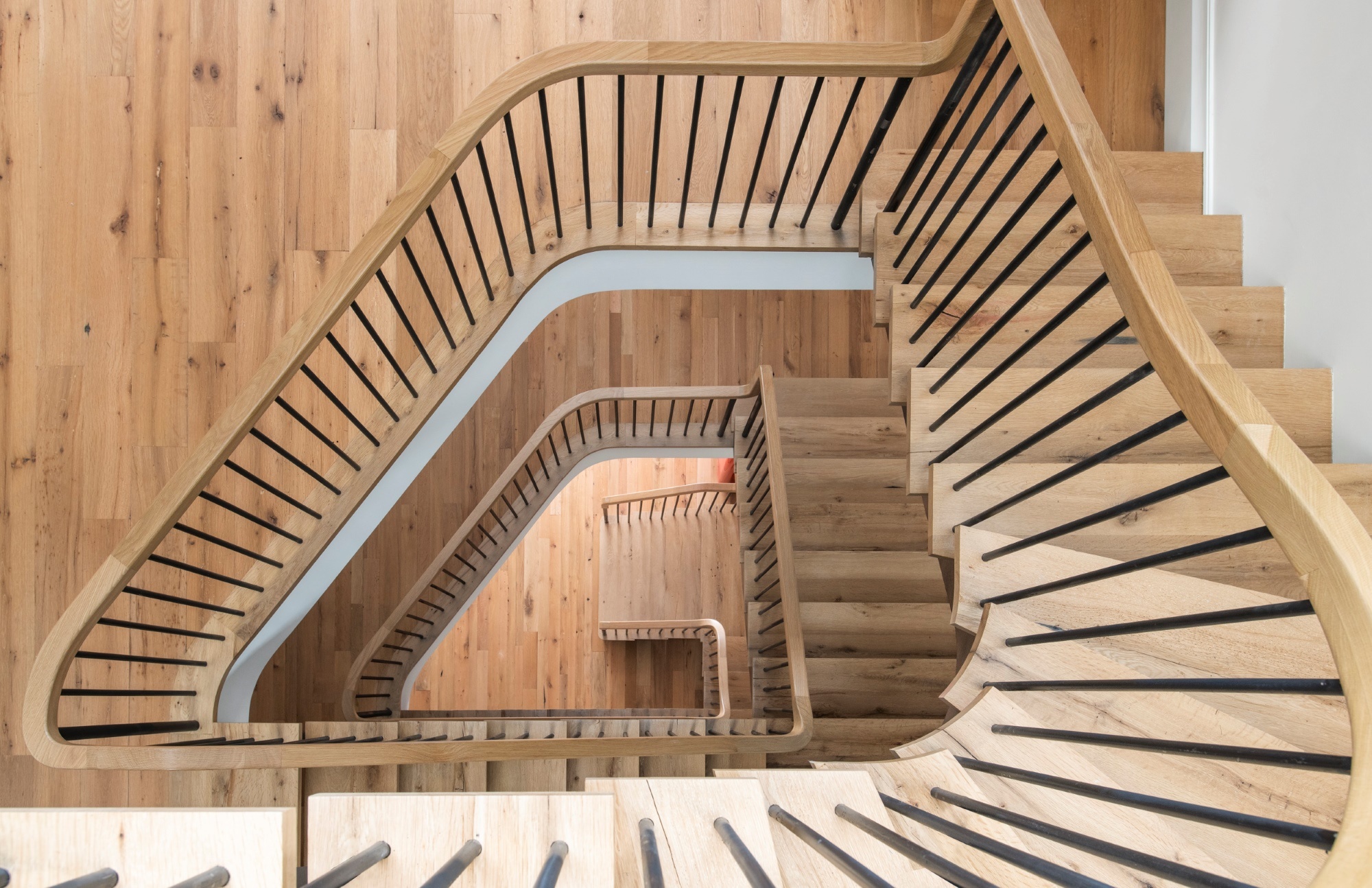 The view from the top of a sculptural spiral staircase. The balusters are modern and black, and the risers and handrail are light wood, as are the wood floors on each level.