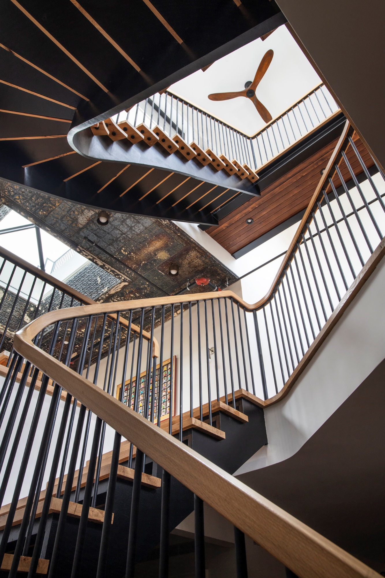 Looking up at a sculptural staircasae in a Manhattan apartment. The staircase is open and modern, and reclaimed tin ceilings can be viewed at the left of the image. A catwalk with wood paneling leads to the outdoor areas, and a modern ceiling fan sits at the ceiling atop the staircase.