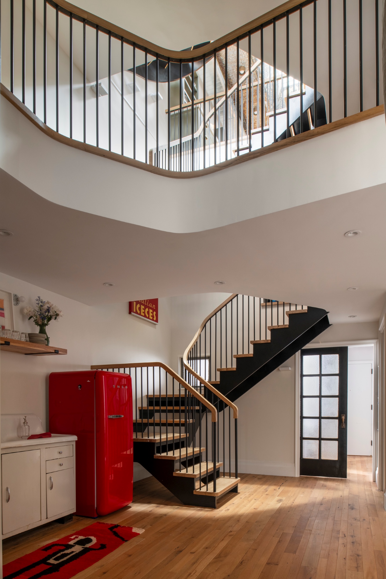 A double height space in a Manhattan apartment. A red SMEG refrigerator sits in the kitchenette next to the landing of the staircase. A lofted space connects the different portions of the fourth floor. Through the open staircase, the top floor kitchen can be seen.