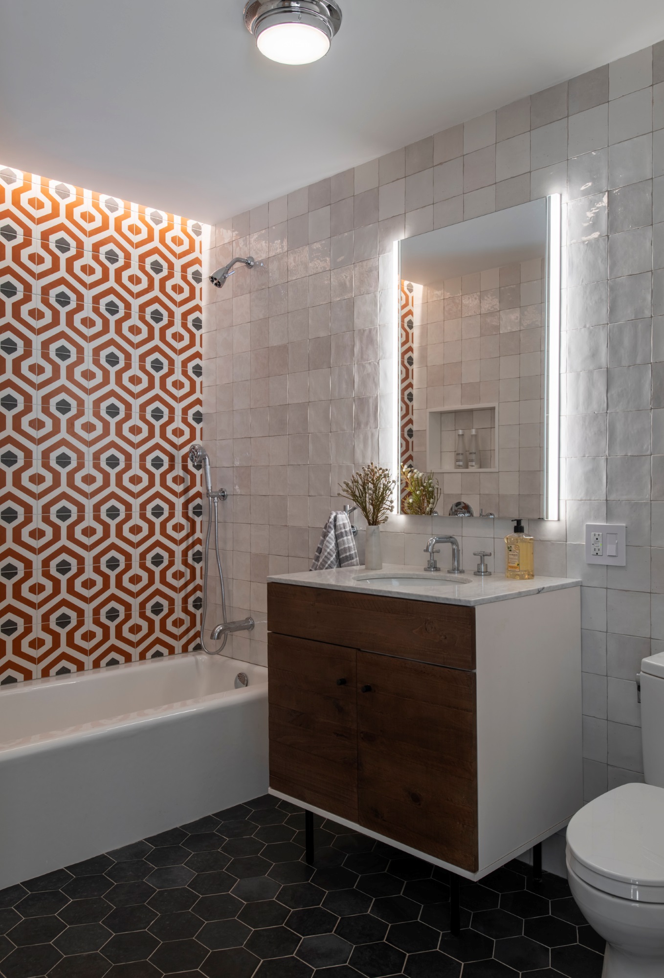 Bathroom in a Manhattan apartment. The floors are dark grey tile, and a floating vanity has dark wood drawers. The walls are tiled, with white tiles along the vanity wall and orange and blue tiles along the tub and shower wall.