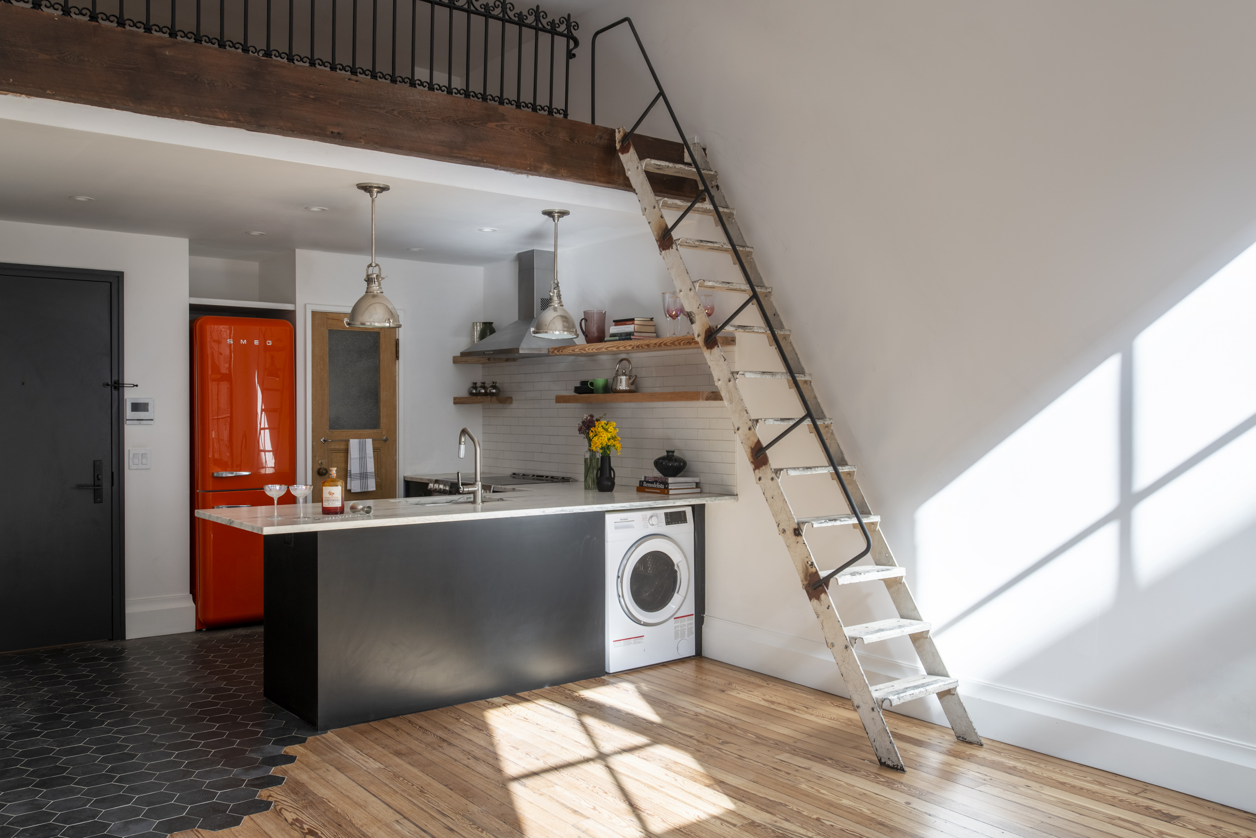 The kitchen and living room space in a Manhattan apartment. The kitchen has a black countertop with white marble counters, an orange SMEG refrigerator, and a reclaimed wood door leading to the pantry. A reclaimed ladder leads to a storage loft above the kitchen.