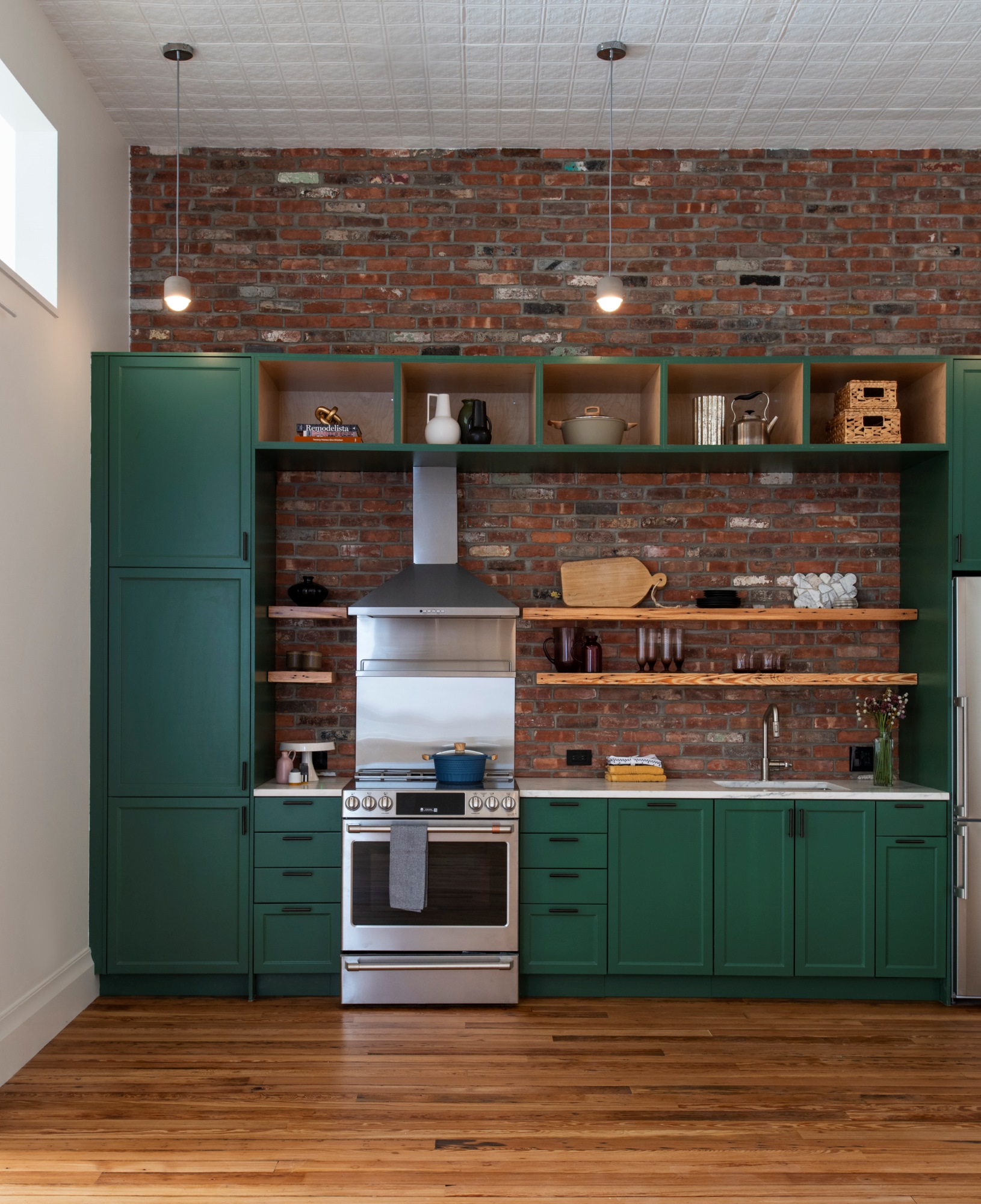 Kitchen in a Manhattan apartment. The wall of the kitchen is exposed brick, the floors are reclaimed wood, and the millwork is painted green. The appliances in the kitchen are all stainless steel, and the ceilings are clad in white painted tin.