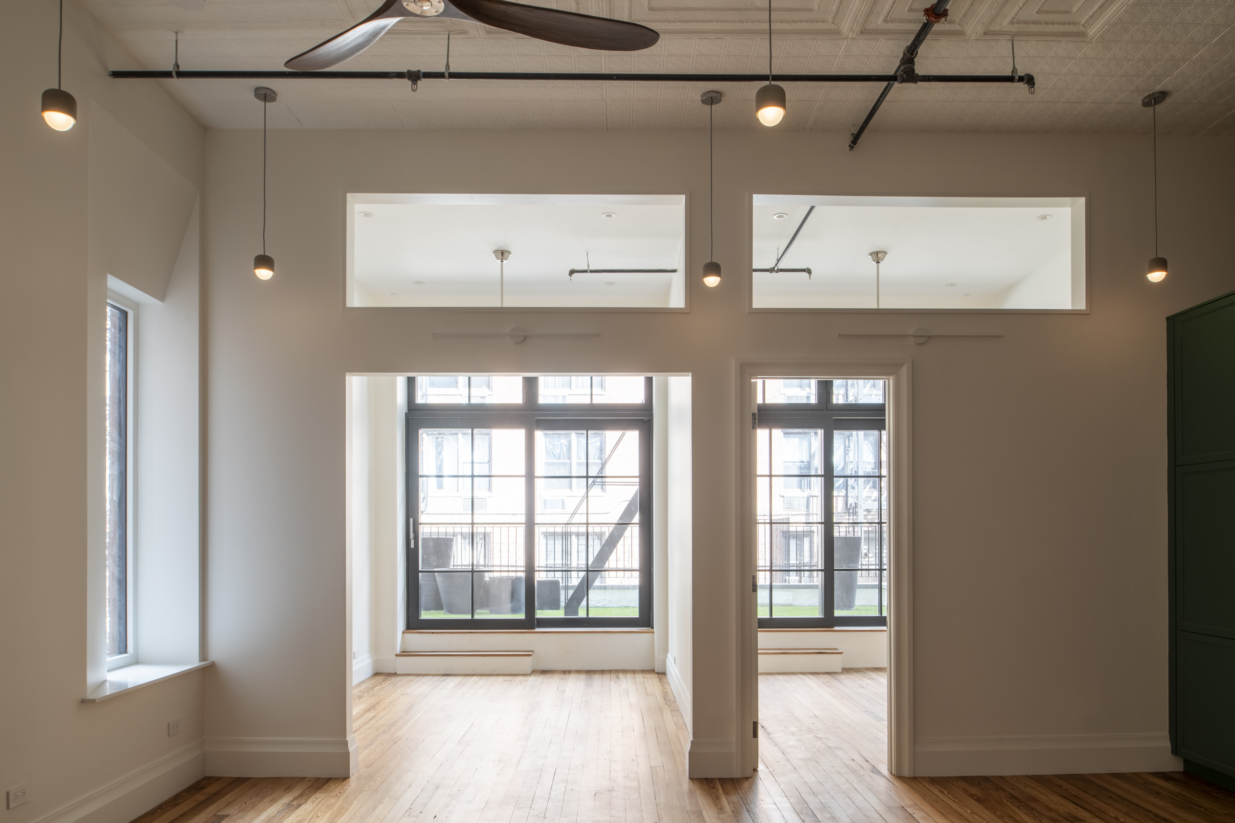 The living room and bedroom in a Manhattan apartment. Large windows lead out to a turfed outdoor area. At the left of the image, a flayed window allows light into the living room.