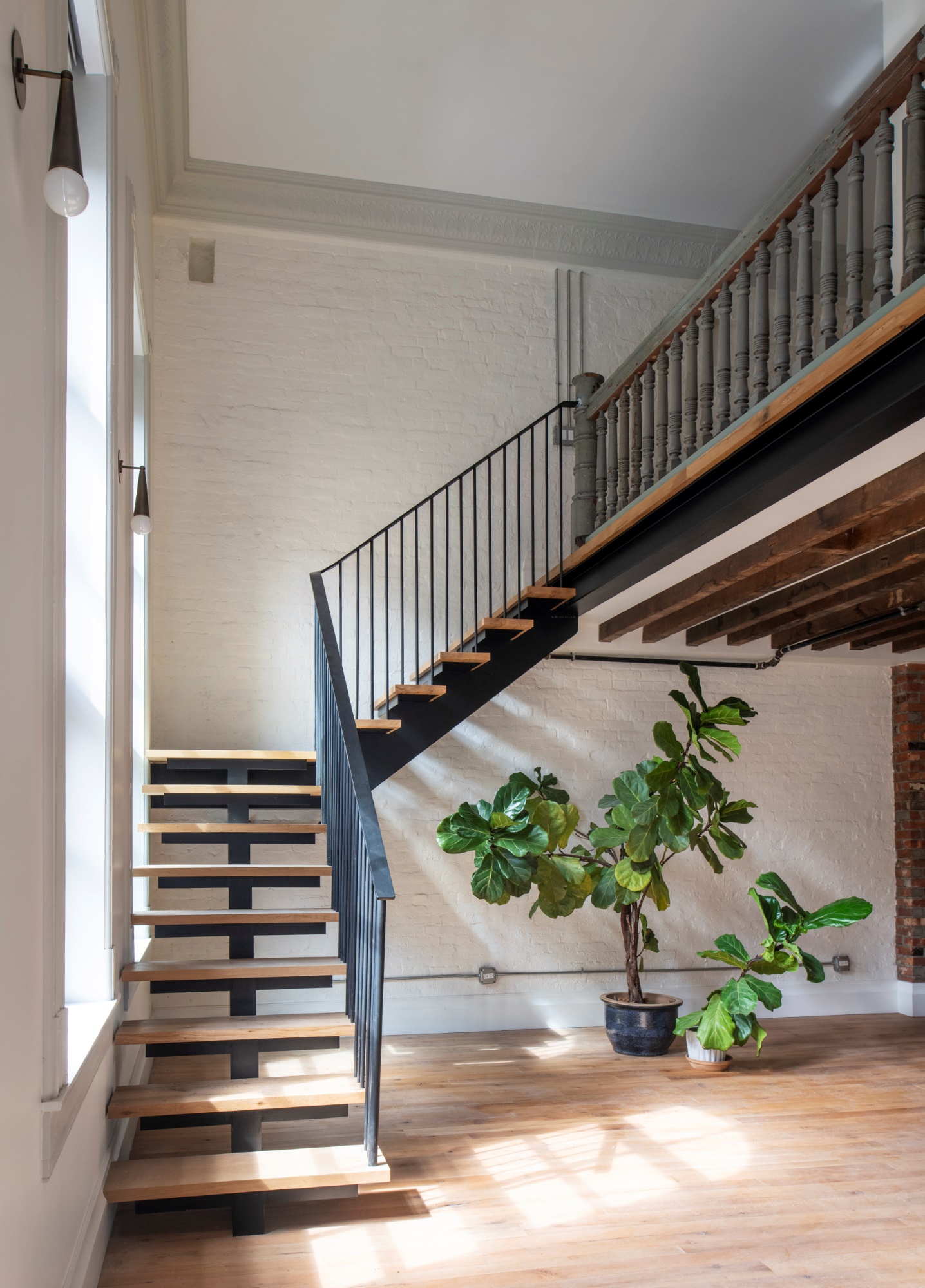 Staircase leading to the lofted bedroom area in a Manhattan apartment. The staircase is modern, with open risers and open stringers. In the living room sits two fiddle leaf fig plants. Reclaimed railings separate the loft from the space below. The walls are white painted brick, and the crown moldings are light grey.