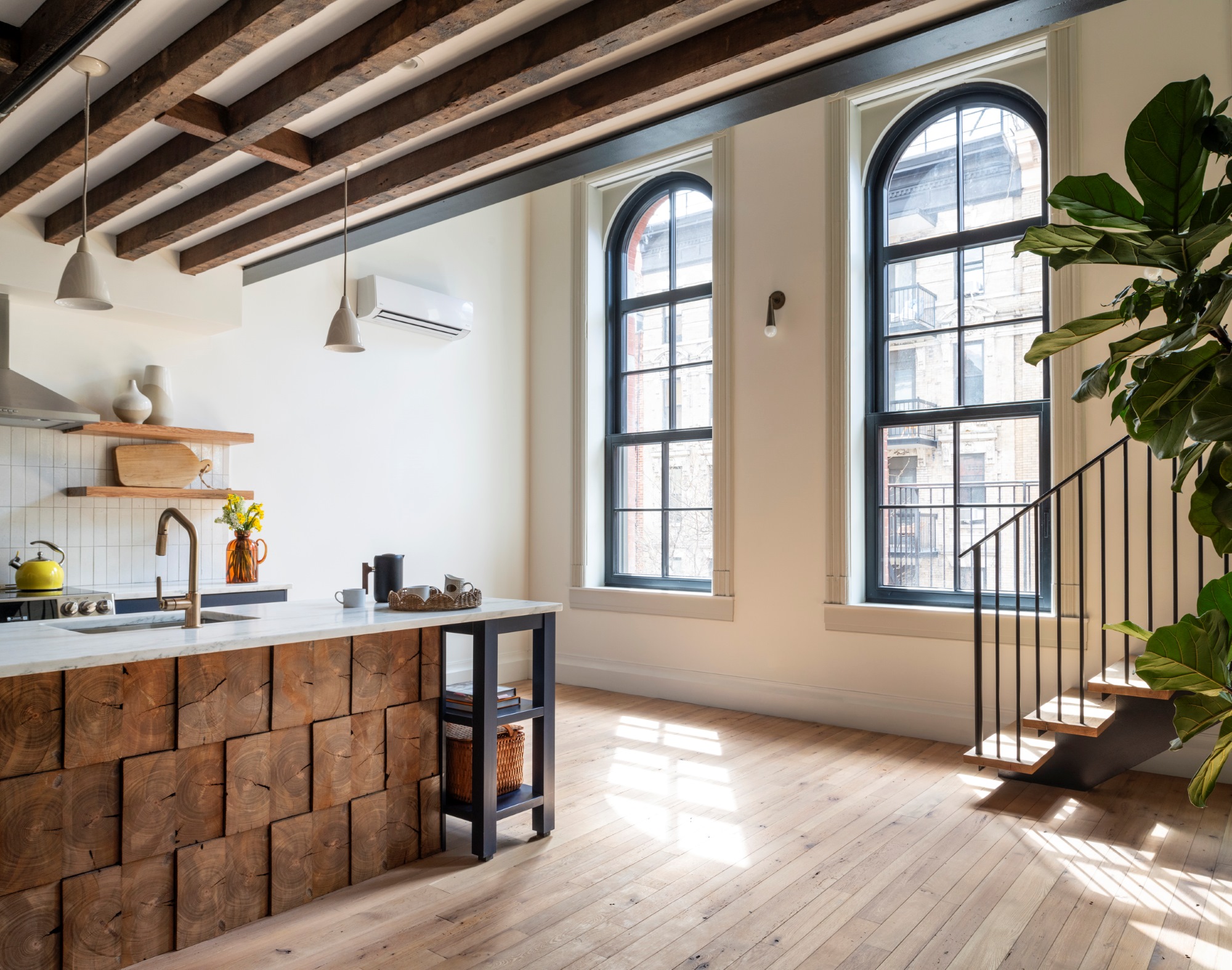 The kitchen at the front of a Manhattan apartment building. The historic window wells have large arched tops. The beams at the ceilings are exposed, and the floors are light reclaimed wood. The large island is clad in reclaimed wood with white marble countertops.