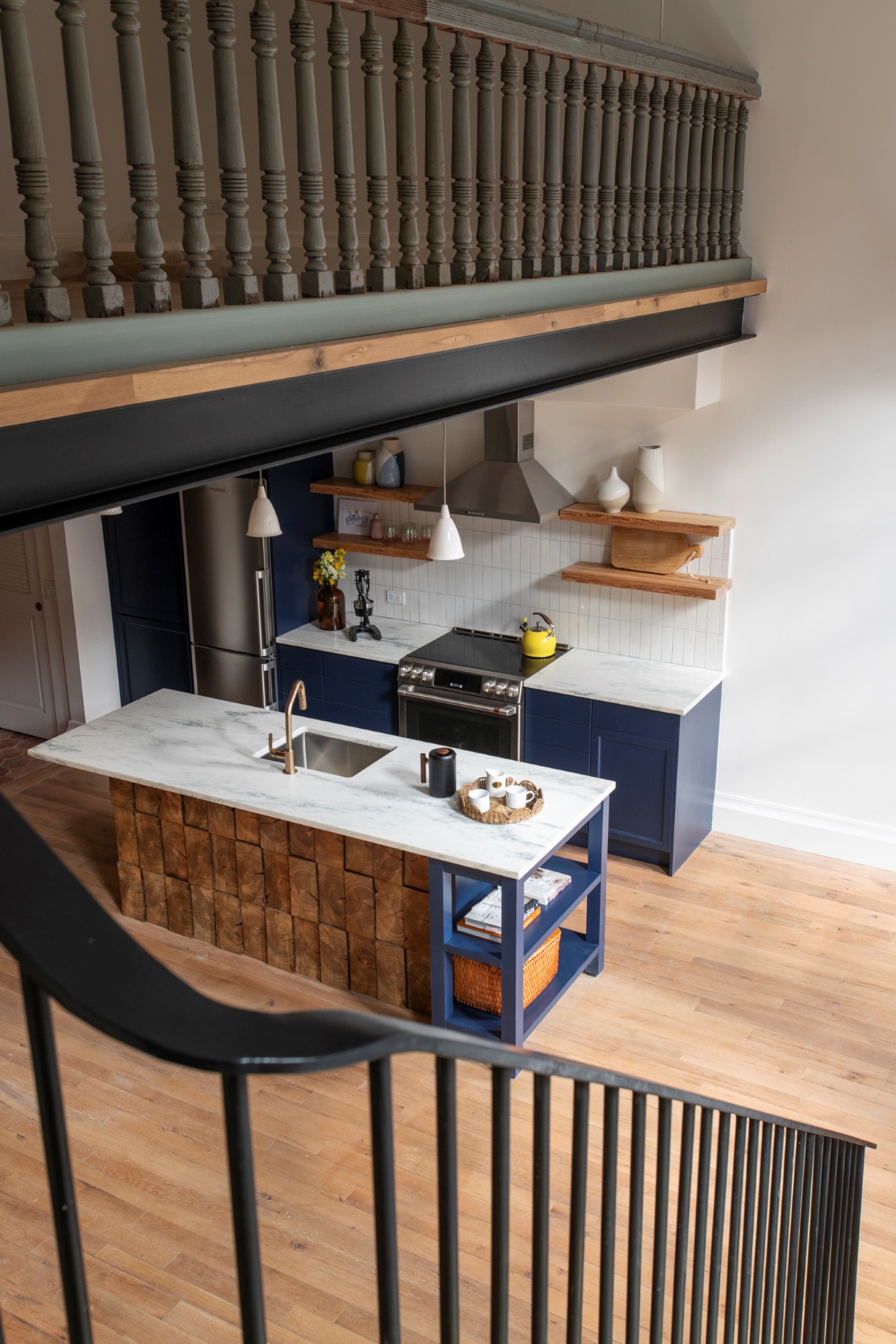 View of the kitchen from the staircase in a Manhattan apartment. At the top of the image, reclaimed railings protect a lofted bedroom area. The kitchen millwork is blue with white marble countertops, and the kitchen island is clad with reclaimed wood from the building's joists.